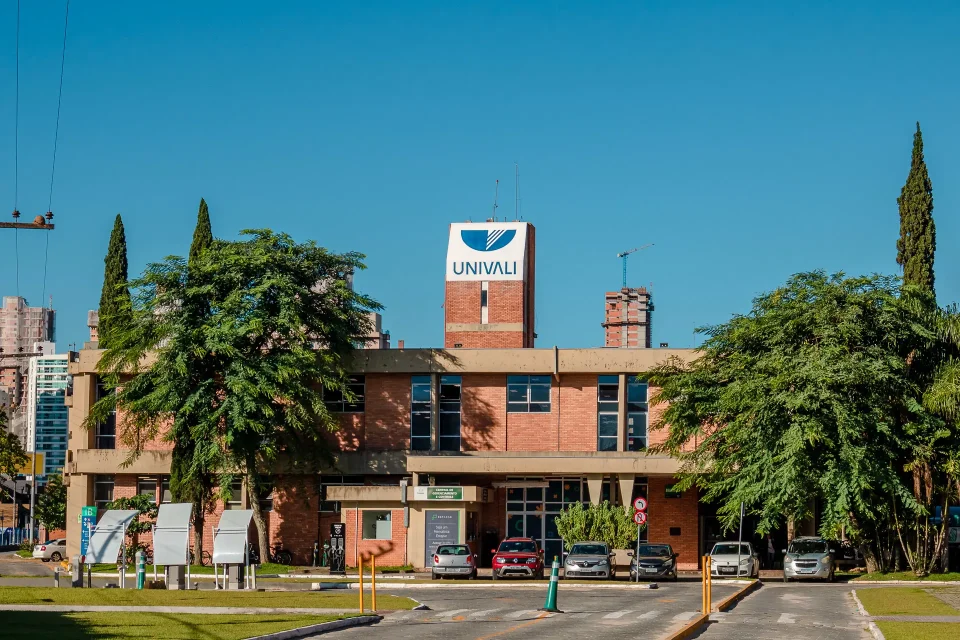 #PraTodosVerem: A foto mostra a fachada de um prédio de tijolos à vista com o letreiro UNIVALI no topo, rodeado por árvores, gramado e carros estacionados.